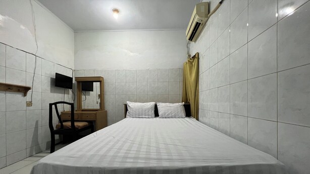 A small, clean room with white tiled walls, a bed with white bedding, a wooden nightstand, a chair, a mirror, and an air conditioning unit. - Hotel O Kost Aisyah Near Citra Grand Galesong City(Pattalassang) (Pattallassang)
