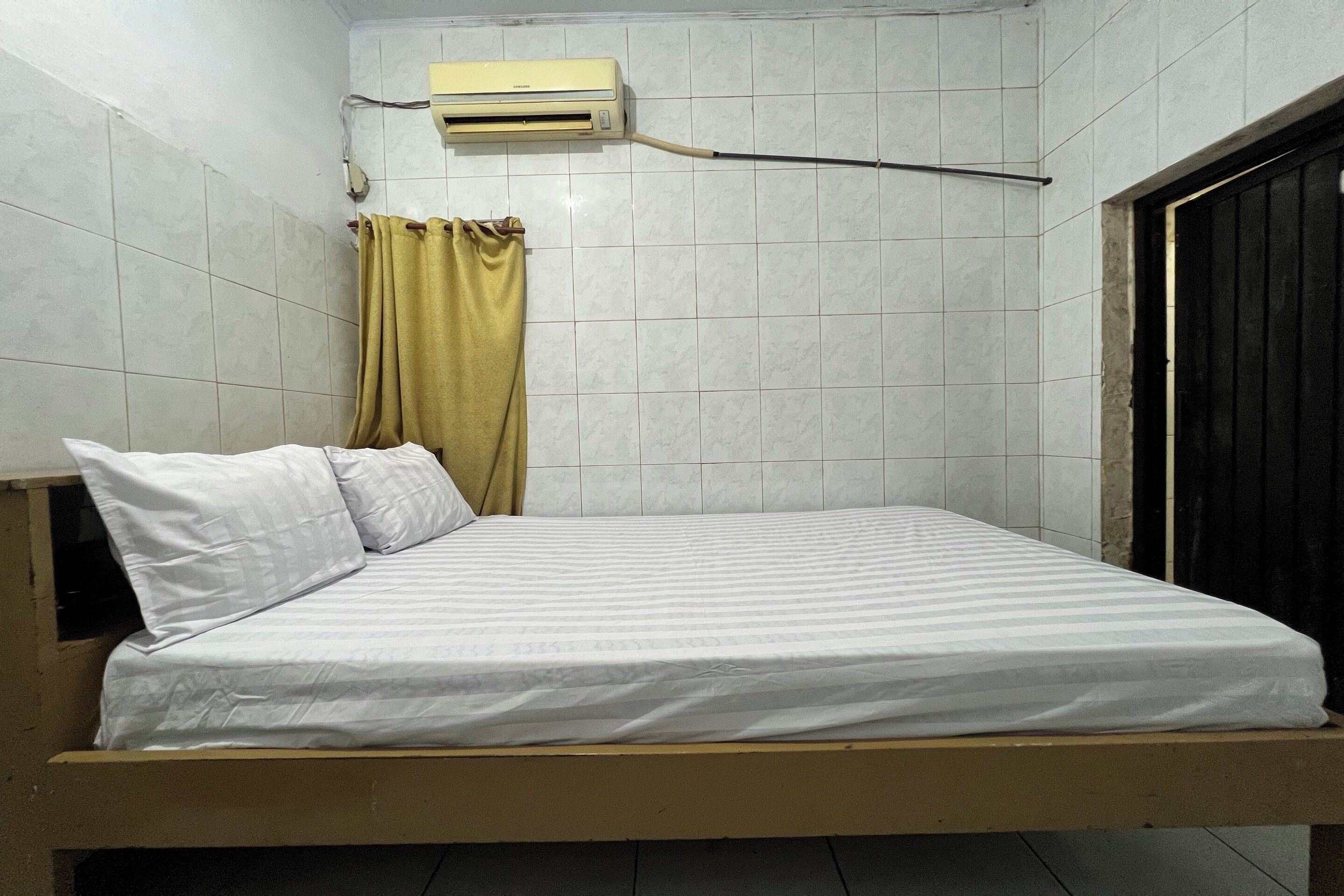 Una cama individual con ropa blanca en una habitación con paredes de baldosas y una ventana con cortina.