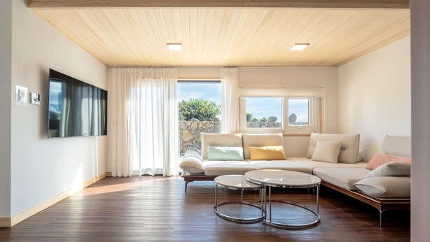 A spacious living room with a wooden ceiling, a large flat-screen TV, a sofa set, a coffee table, and a view of the outdoors through large windows. - Jeju Staysosoheon Dokchae Pension (Jeju City)