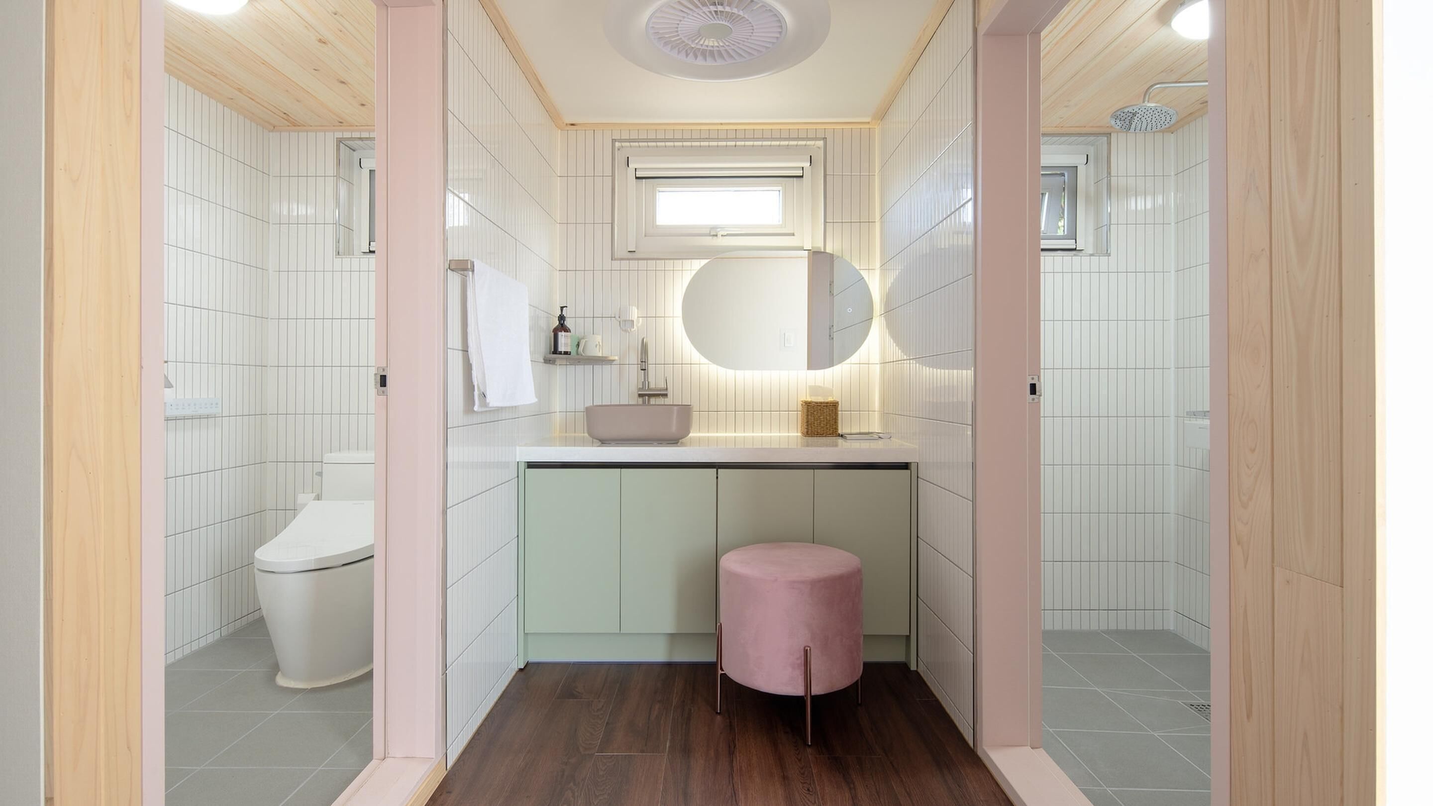A compact bathroom with a toilet, sink, and vanity, featuring a round mirror, wooden flooring, and a small stool.