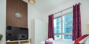 A modern hotel room with a flat-screen TV, a large window with curtains, a bed with pillows, and a wardrobe. - 360 Vacation - Beach Side Classic Studio (Dubai)