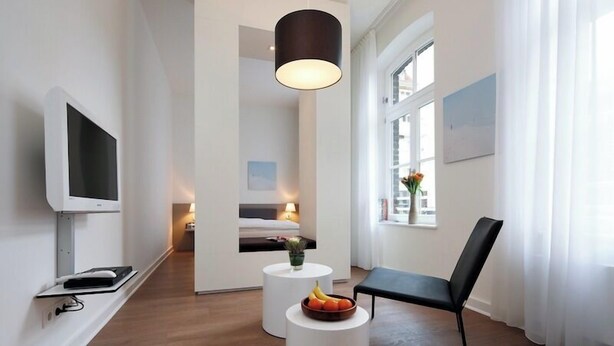Ett modernt hotellrum med en platt-TV, ett runt soffbord med frukt på, en svart stol och ett stort fönster med gardiner.