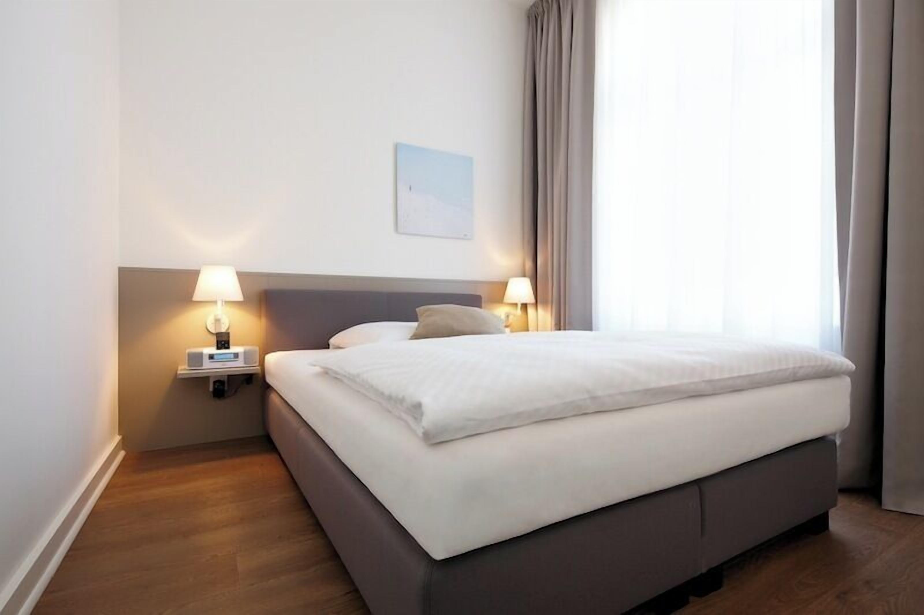 Ein ordentlich bezogenes Bett mit weißen Leinen, zwei Nachttischlampen, ein kleiner Nachttisch und ein an der Wand hängendes Bild.