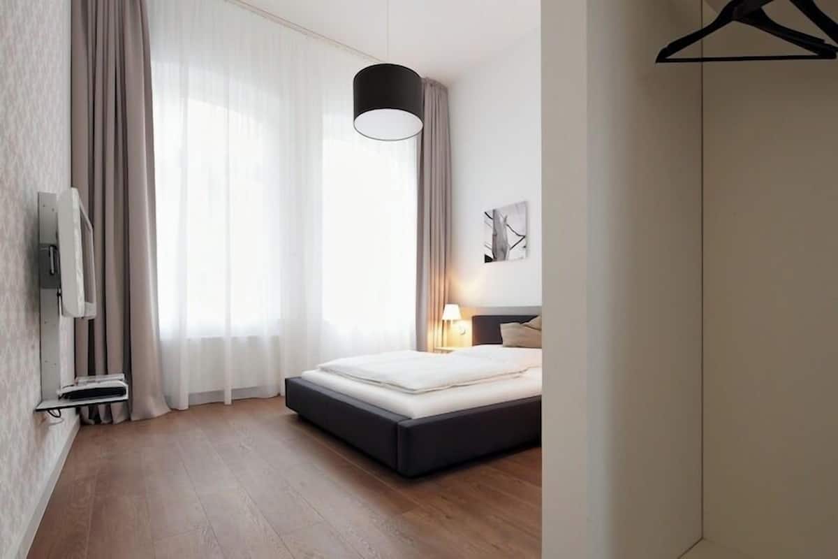 A modern hotel room with a bed, a hanging light fixture, a wall-mounted TV, and a window with curtains.