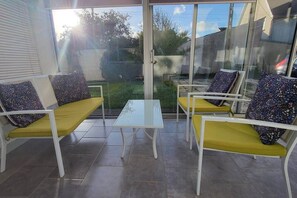 A sunlit patio with white metal chairs and yellow cushions, a small glass table, and a view of a garden and street.