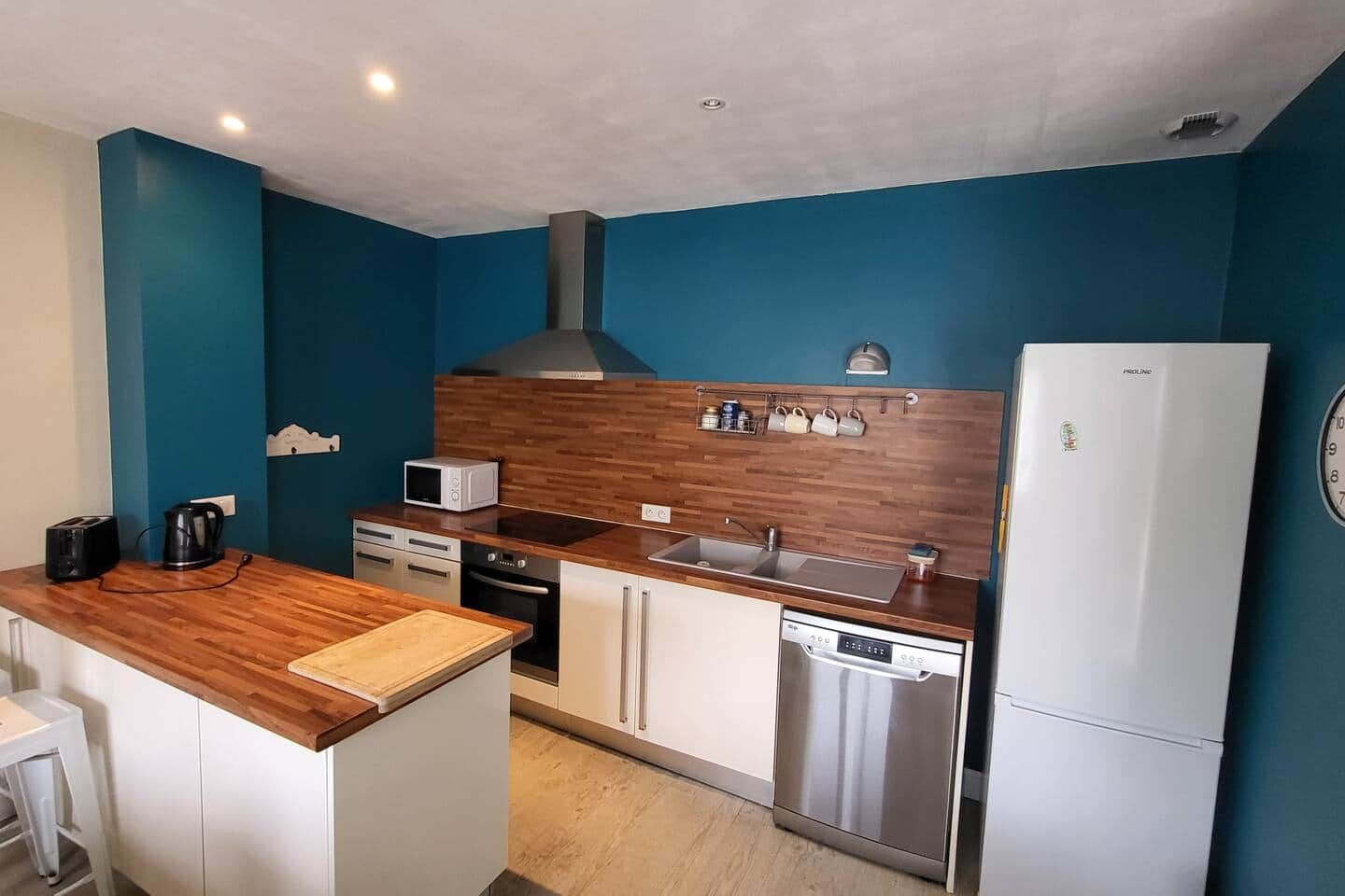 Une cuisine compacte dotée d’un plan de travail en bois, de meubles blancs, d’un four à micro-ondes encastré, d’un lave-vaisselle en acier inoxydable et d’un réfrigérateur blanc.