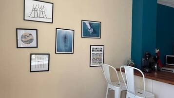 Une pièce avec un mur de couleur beige, un mur d’accent bleu et une chaise blanche. Des œuvres d’art encadrées ornent le mur, et une machine à café est posée sur le comptoir.