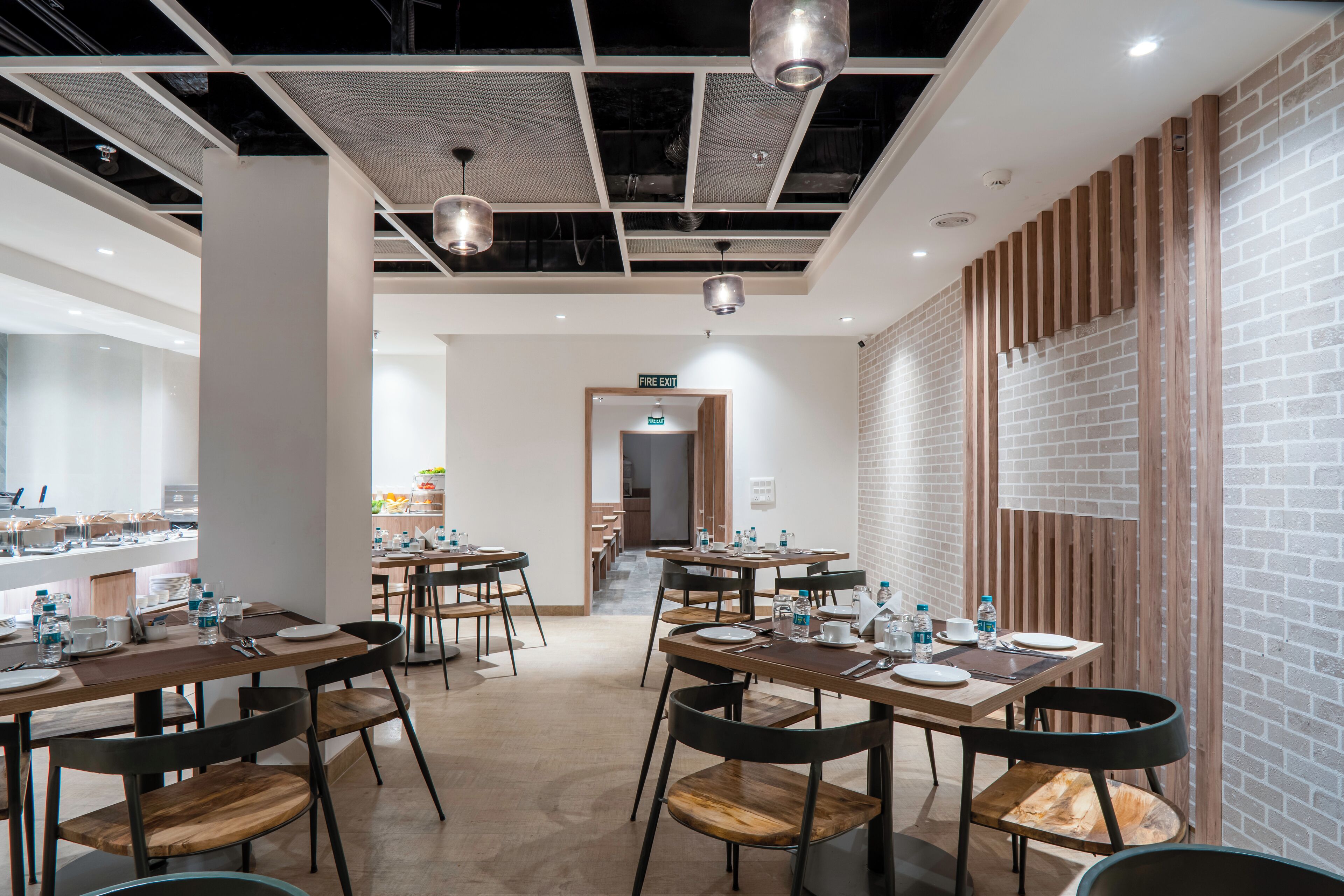 Un comedor moderno con varias mesas y sillas, un letrero de salida de incendios visible y una pared de ladrillo en un lateral.