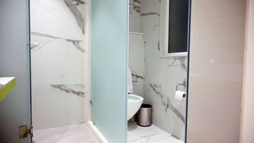 A bathroom with a marble floor, a toilet, a trash bin, and a glass shower enclosure.