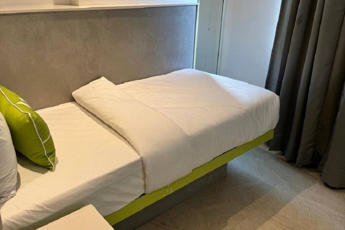 A single bed with a green frame in a room with a plain white wall and a grey curtain.