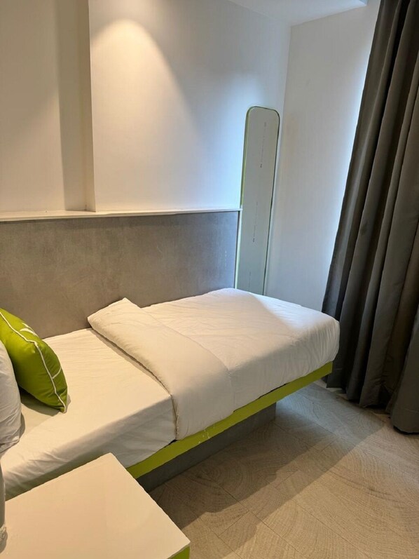 Una cama individual con estructura verde en una habitación con pared blanca lisa y cortina gris.