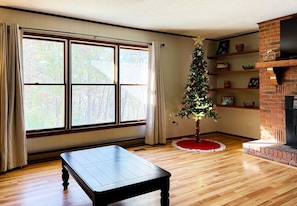 Um quarto com piso de madeira, lareira, árvore de Natal e uma grande janela com cortinas.