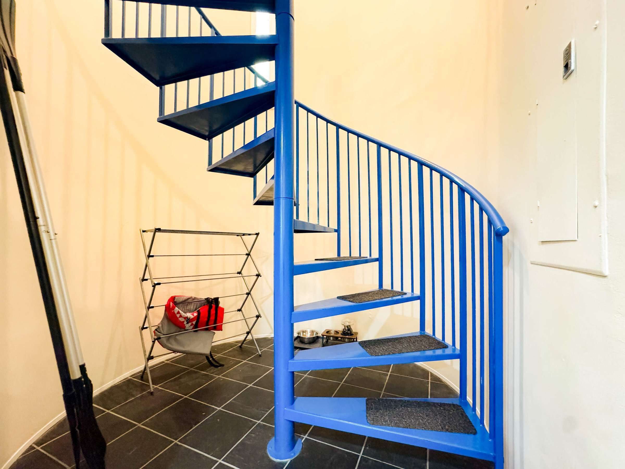 A blue spiral staircase in a room with tiled flooring and a metal drying rack with a red bag.