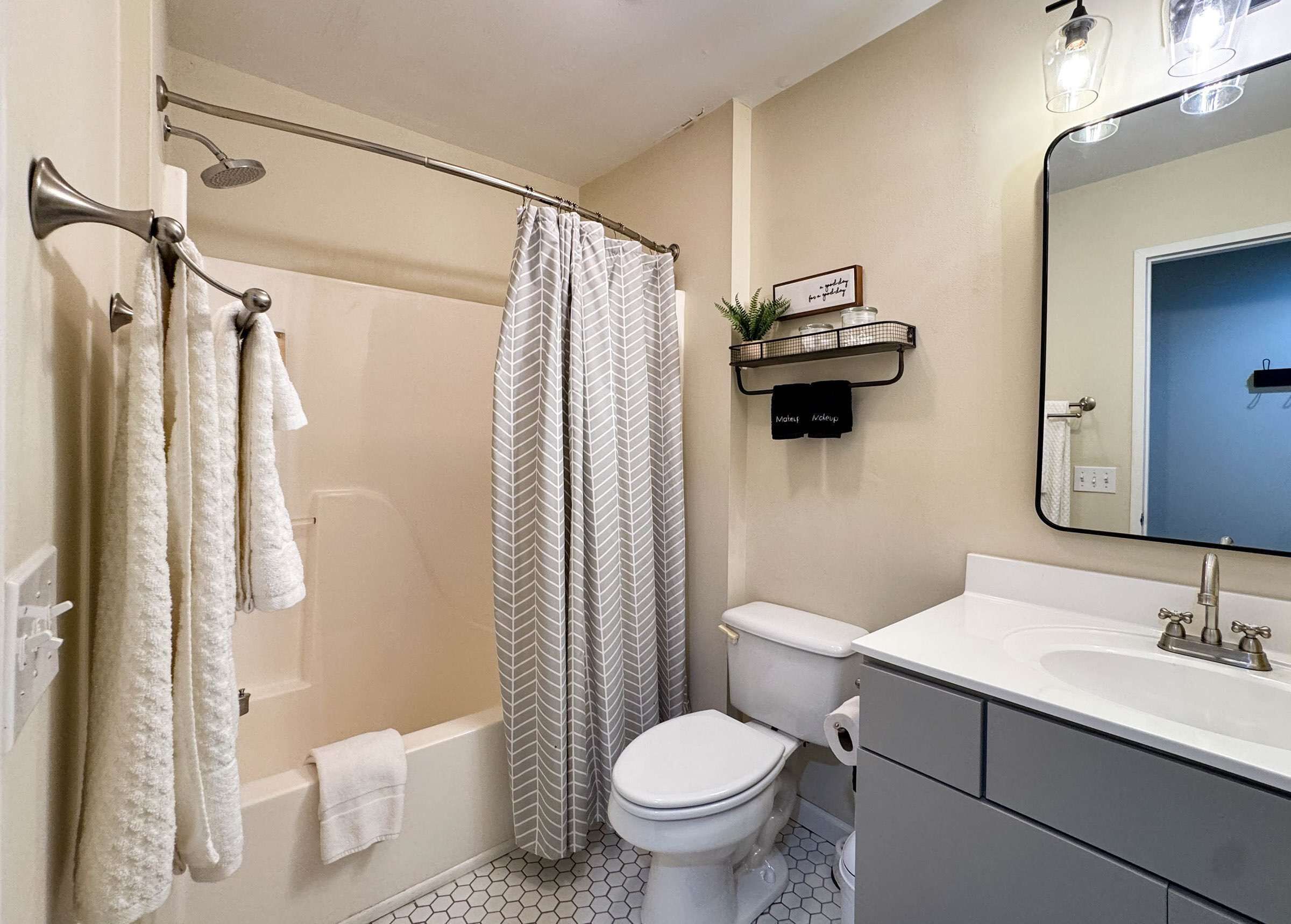 A bathroom with a shower, toilet, and sink. There is a towel rack with towels, a shower curtain, and a small shelf with a plant.
