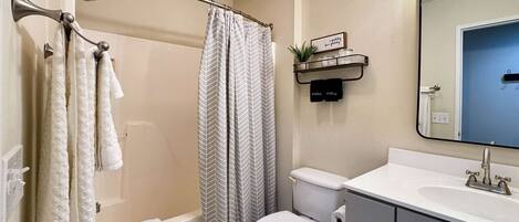 A bathroom with a shower, toilet, and sink. There is a towel rack with towels, a shower curtain, and a small shelf with a plant.