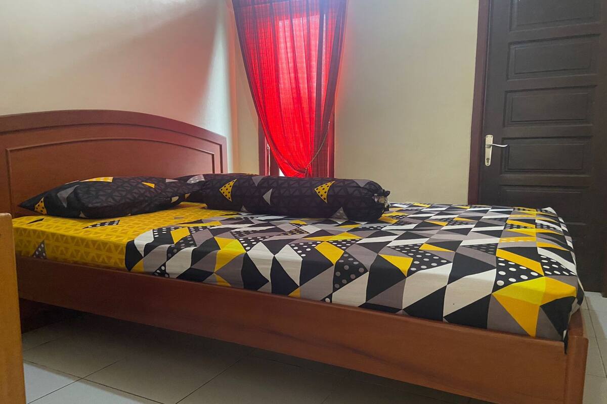 Una cama con una colcha y almohadas de estampado geométrico, un cabecero de madera, una cortina roja y una puerta cerrada.