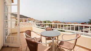 Ein offener Balkon mit Korbstühlen und einem Tisch, der Blick auf eine Küstenstadt und das Meer gewährt.