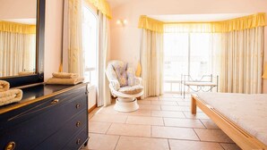 Ein Hotelzimmer mit Bett, Stuhl, Kommode und einem großen Fenster mit Vorhängen.