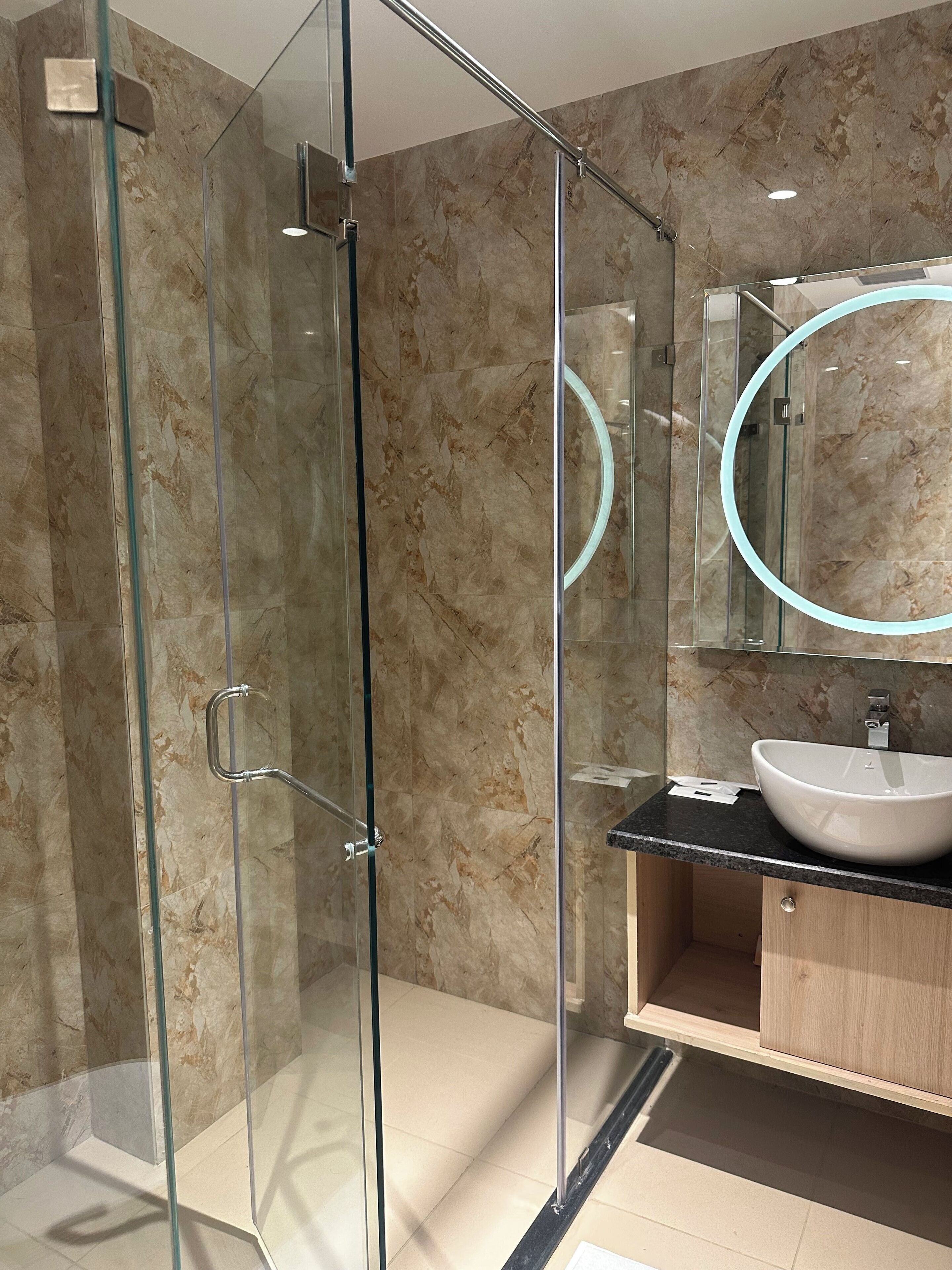 Un baño moderno con ducha de vidrio, pared revestida de mármol, tocador de madera y espejo redondo con luz.
