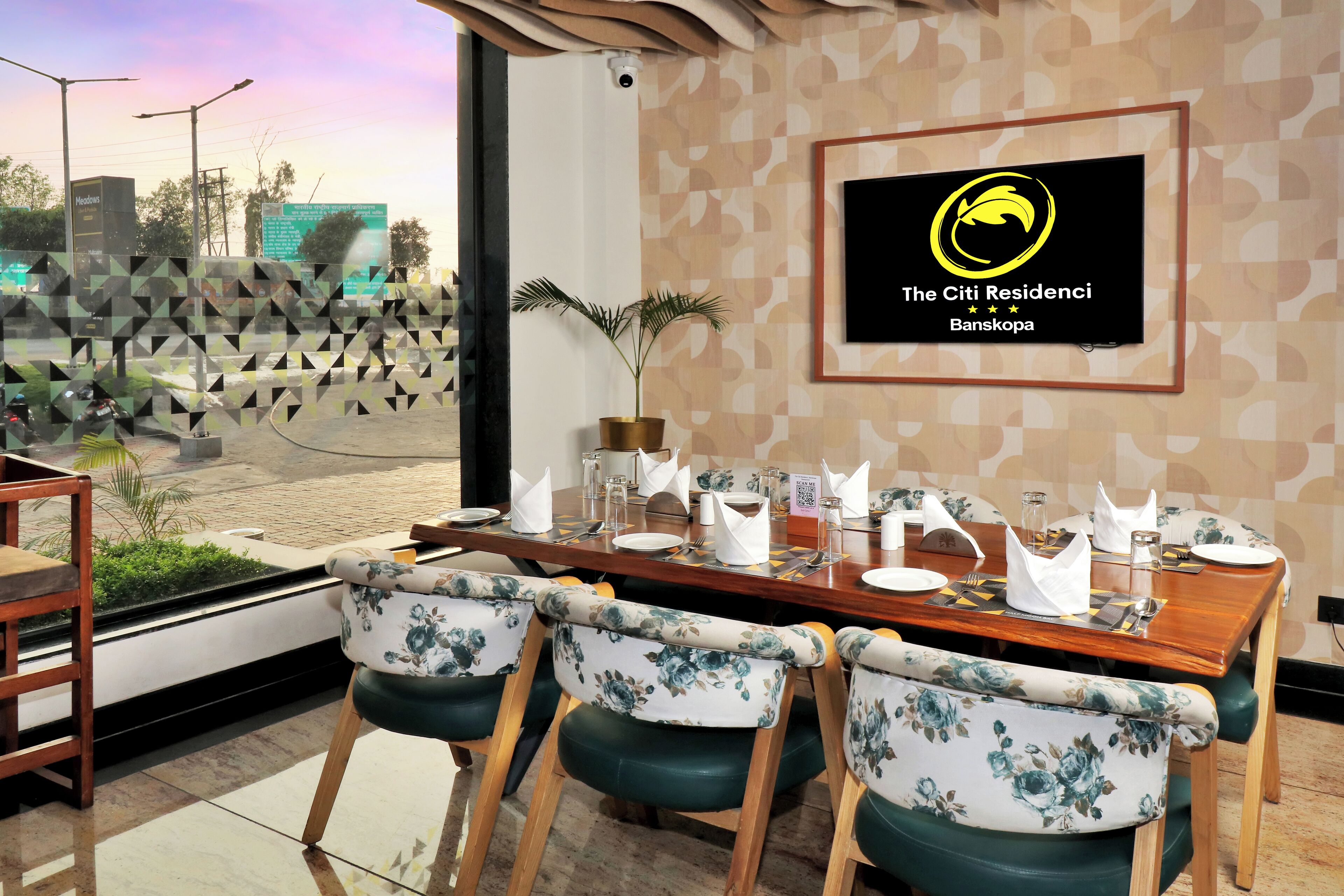 A dining area with a wooden table set for four, surrounded by floral-patterned chairs. A potted plant is on the table, and a framed logo is on the wall.