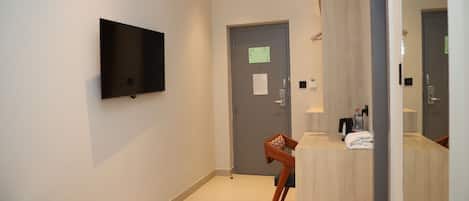 A hotel room with a bed, a TV mounted on the wall, a wooden chair, a desk with a mirror, and a door with a notice.