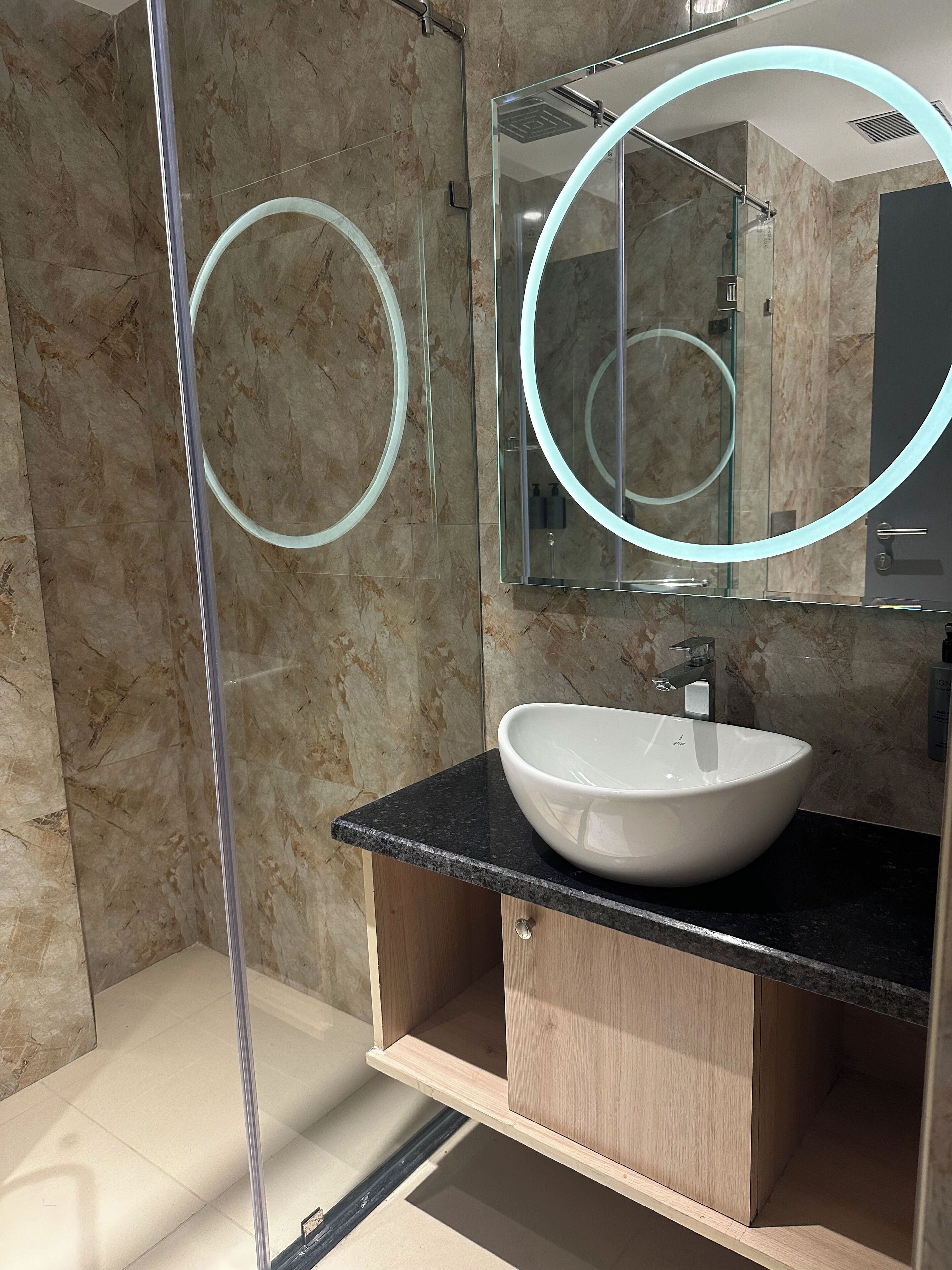 A modern bathroom with a marble wall, a glass shower enclosure, a white sink, and a mirror with a circular frame.