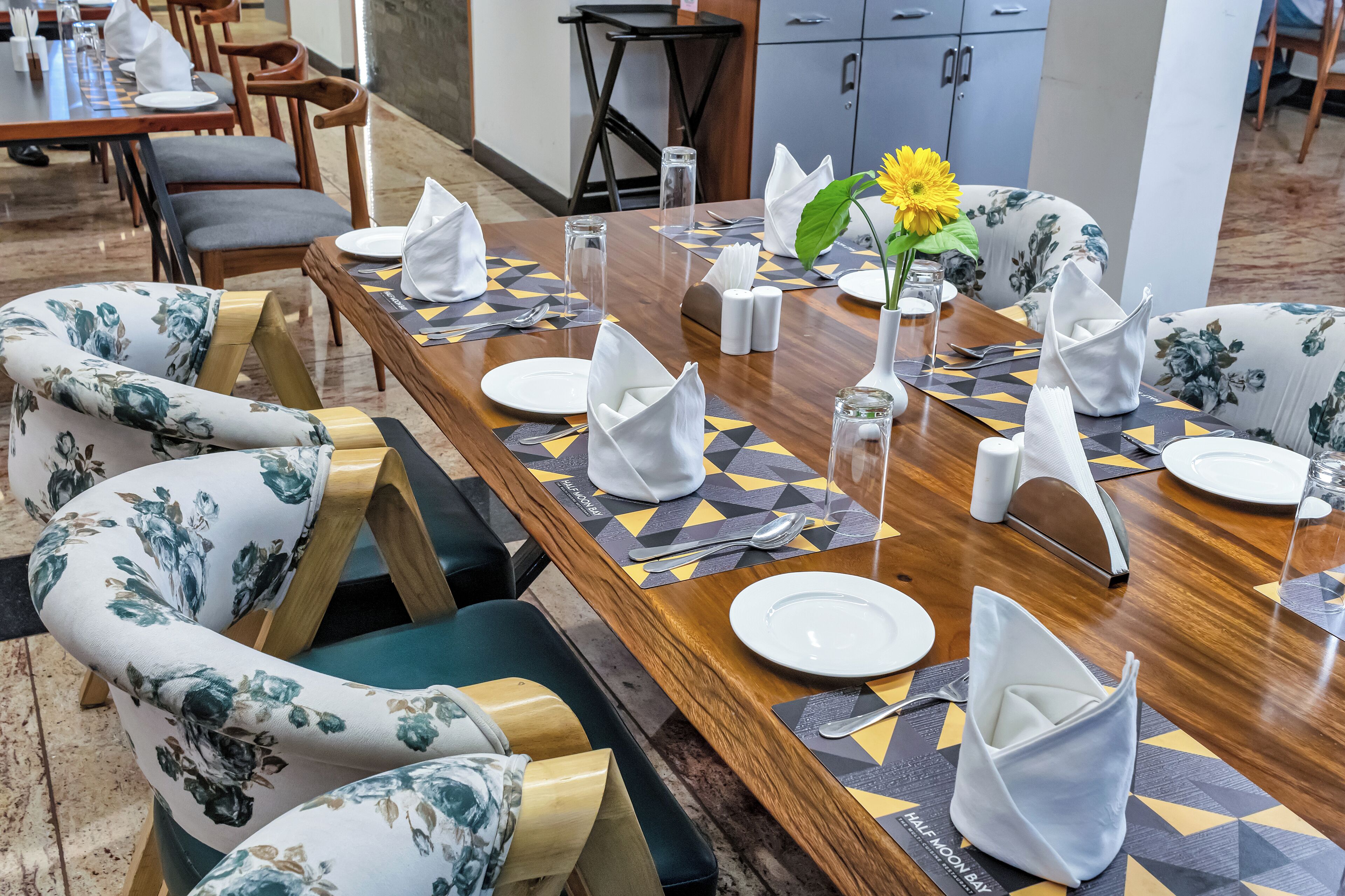 A dining area with a long wooden table set for multiple guests, featuring geometric-patterned placemats, white napkins, and floral-patterned chairs.