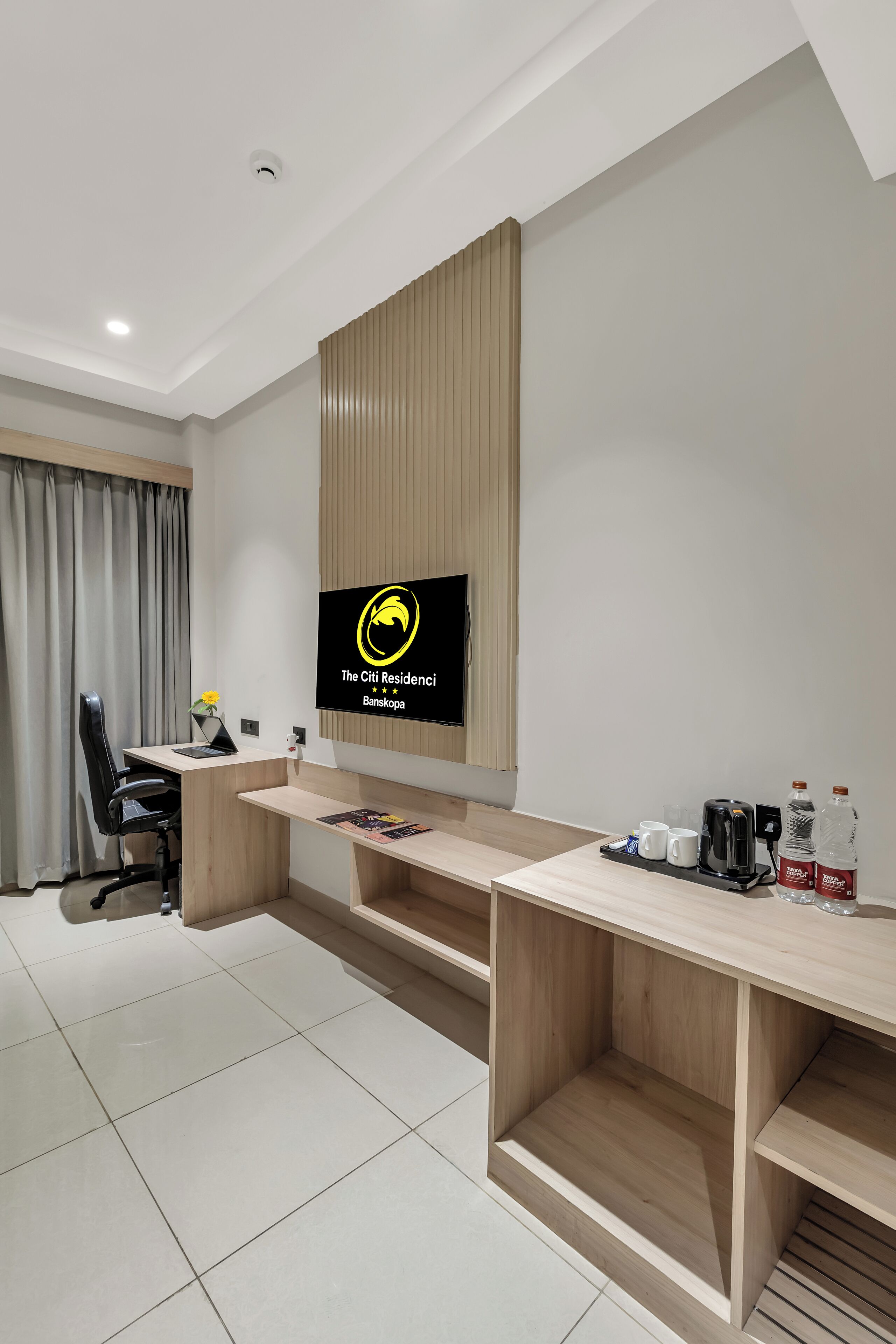 A modern hotel room with a wooden desk, a flat-screen TV mounted on the wall, a coffee machine, and a small shelf with cups and bottles.