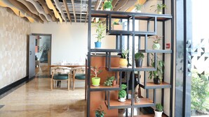 Un intérieur moderne avec un plafond en bois, une étagère en métal noir agrémentée de plantes, un coin repas avec une table et des chaises, et un mur orné d’un motif.