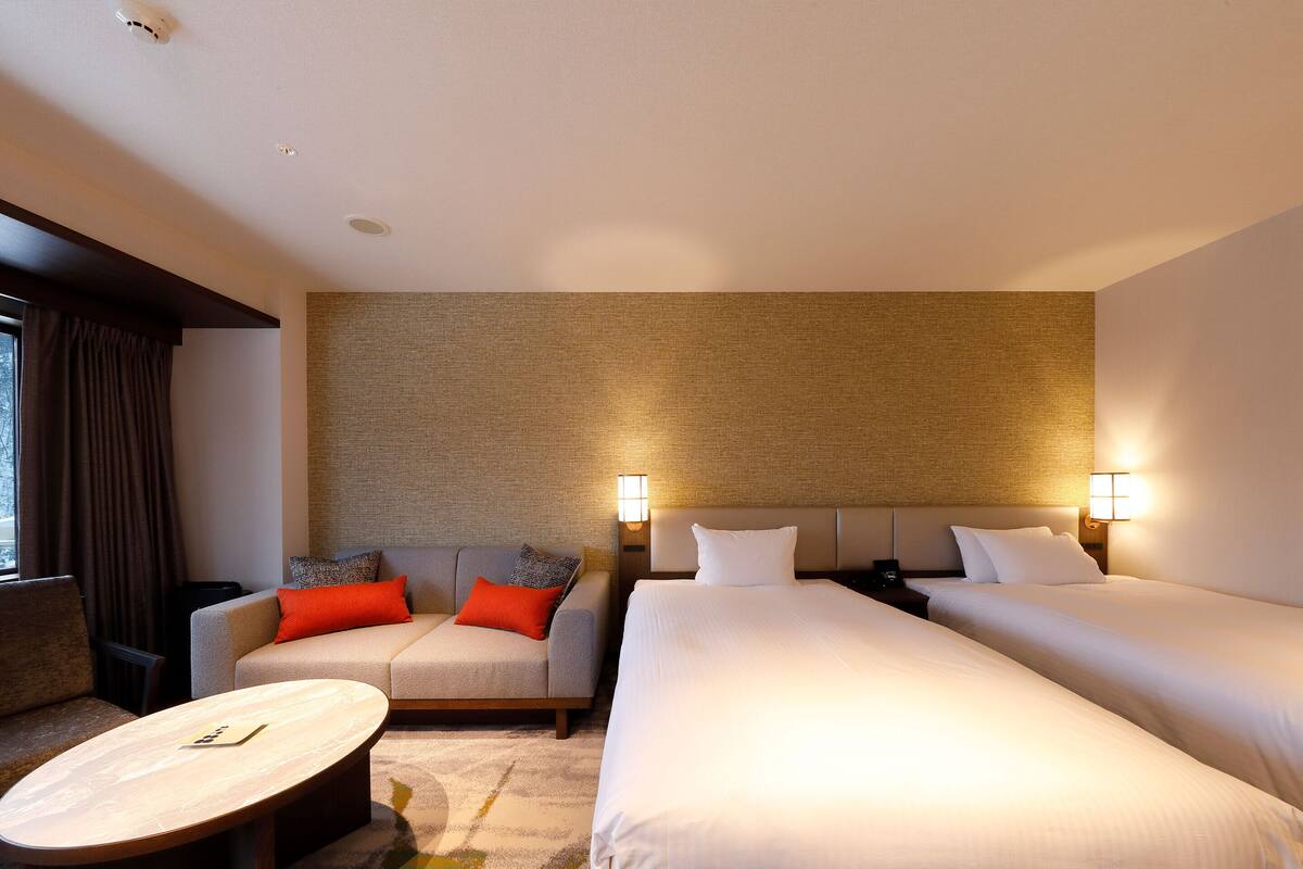 Habitación de hotel con dos camas, un sofá, una mesita y una lámpara fijada a la pared.