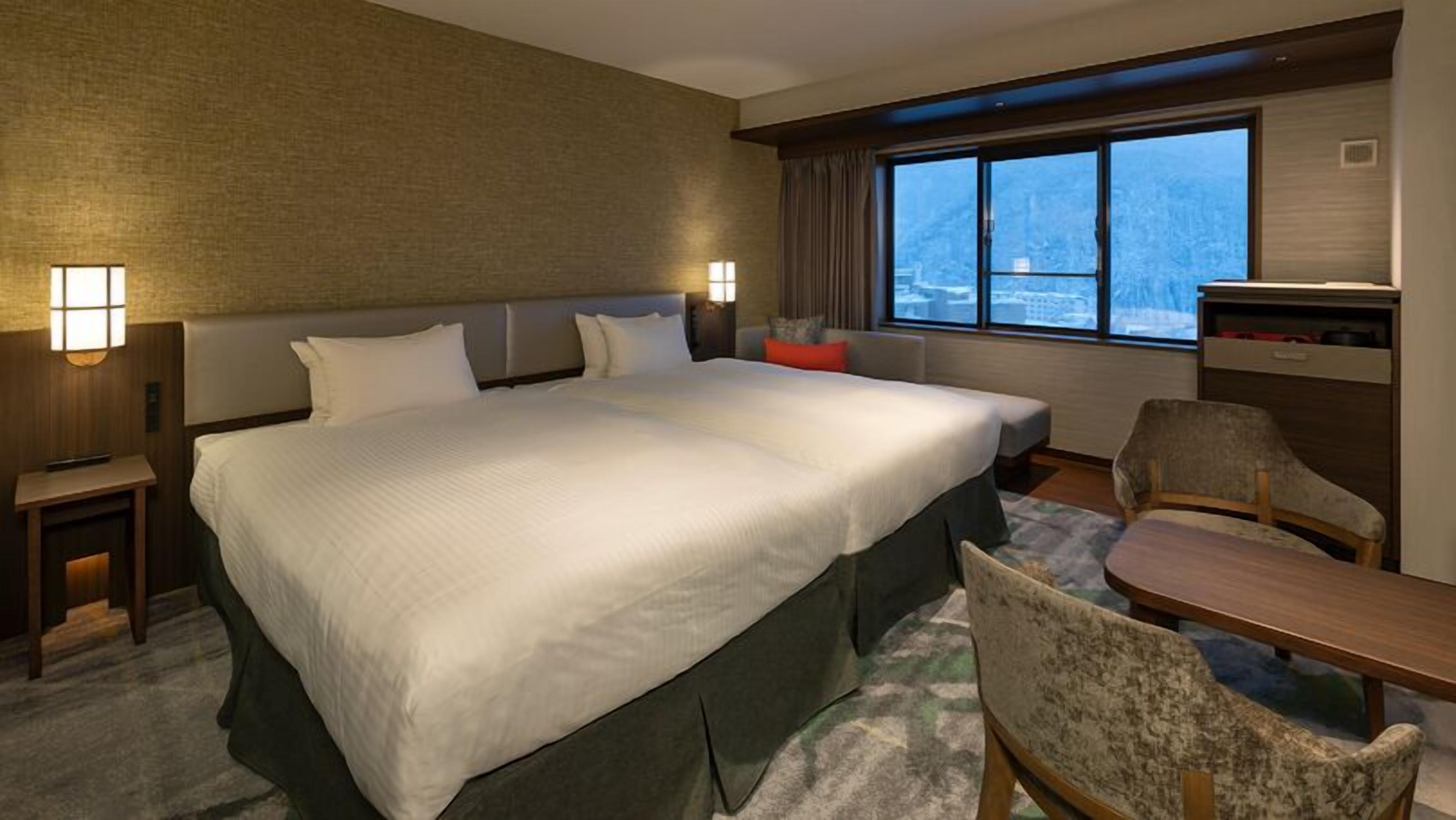 A hotel room with a large bed, a desk, and a chair, a window with a city view, and a wall-mounted TV.