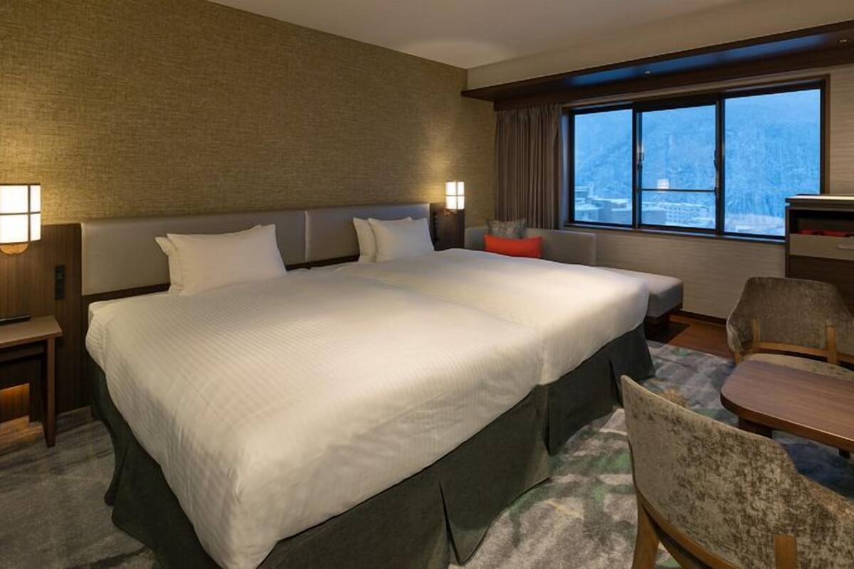 Habitación de hotel con una cama grande, un escritorio, una silla, un ventanal con vista a la ciudad y un televisor montado en la pared.