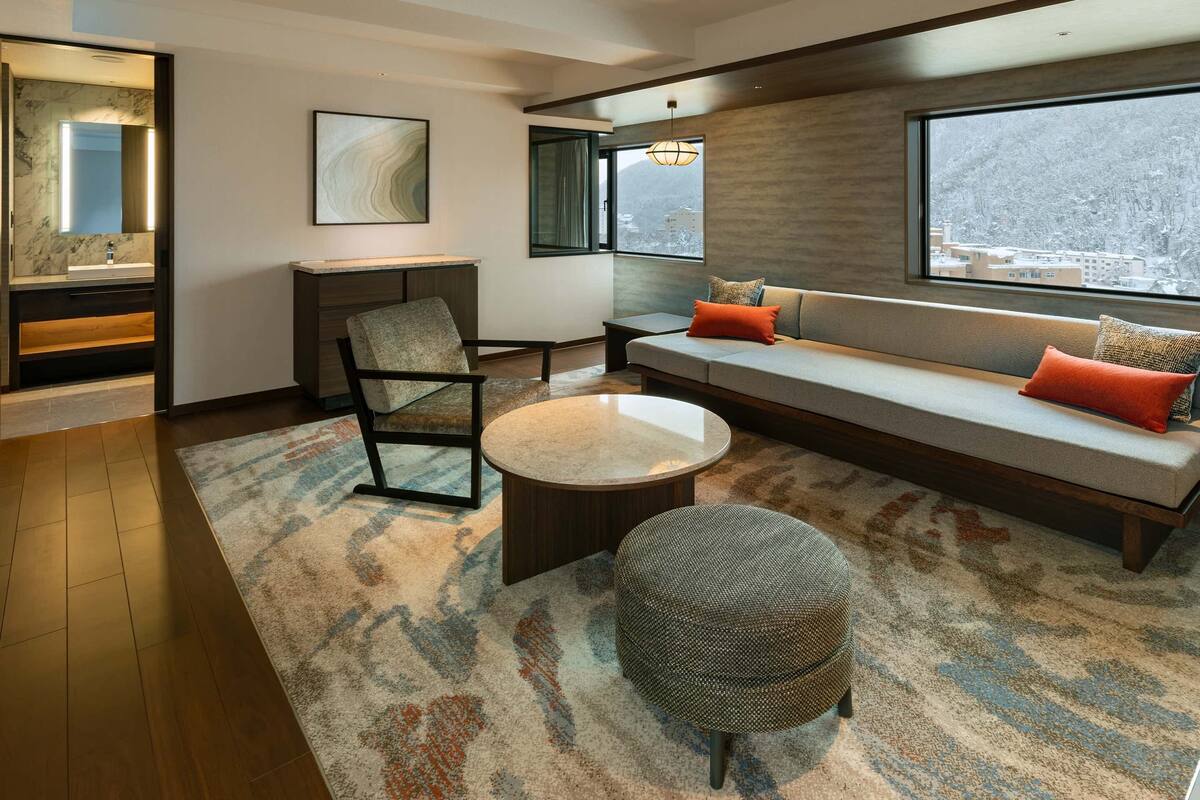 Una sala de estar moderna con un sofá, un reposapiés y una silla, una mesa de centro redonda, un ventanal con vistas y una zona con suelo de mármol y un lavabo.