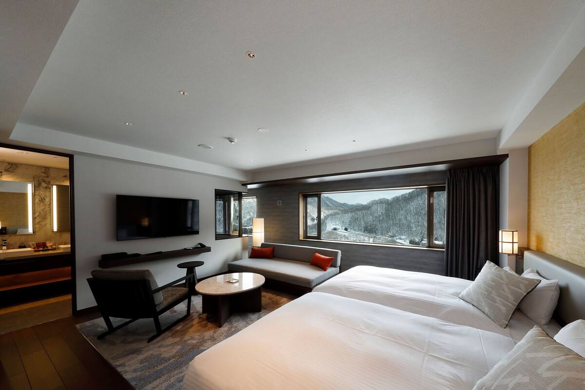 Habitación de hotel con una cama grande, un sofá, un escritorio con silla, un televisor y una ventana con vistas a la montaña.