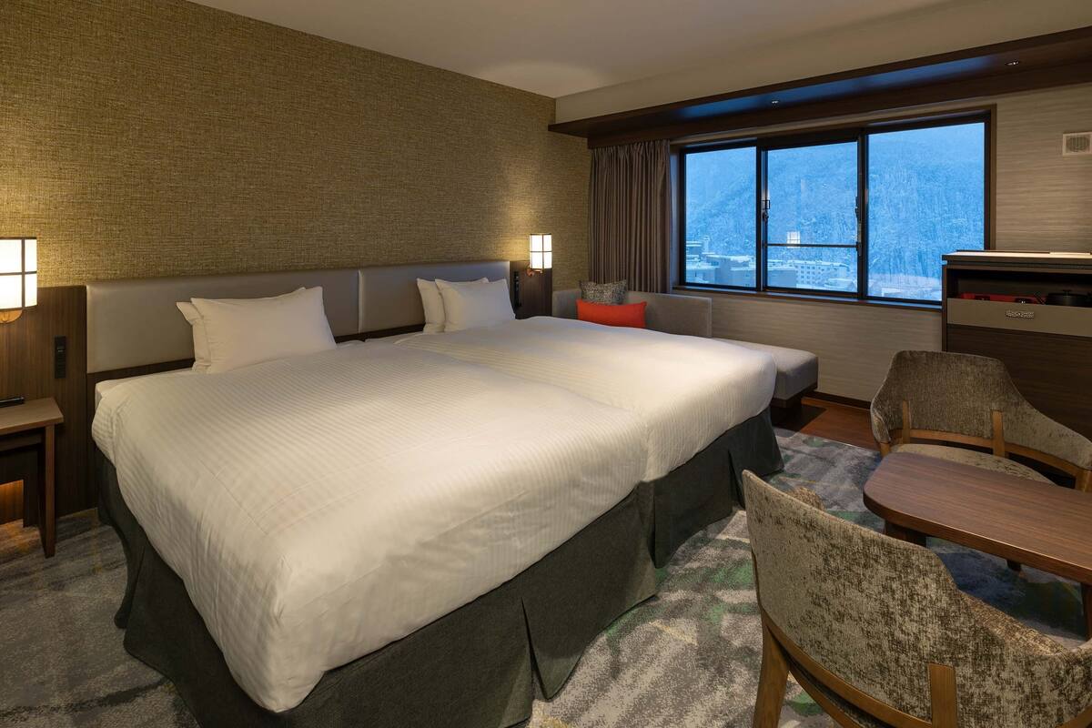 Habitación de hotel con una cama grande, un escritorio, una silla y vista a un paisaje nevado a través de la ventana.