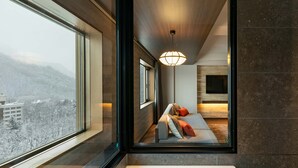 A modern bathroom with a large bathtub, a sofa with cushions, and a view of a snowy cityscape through a large window. - EXECUTIVE SUITE SUIREI (Sapporo)