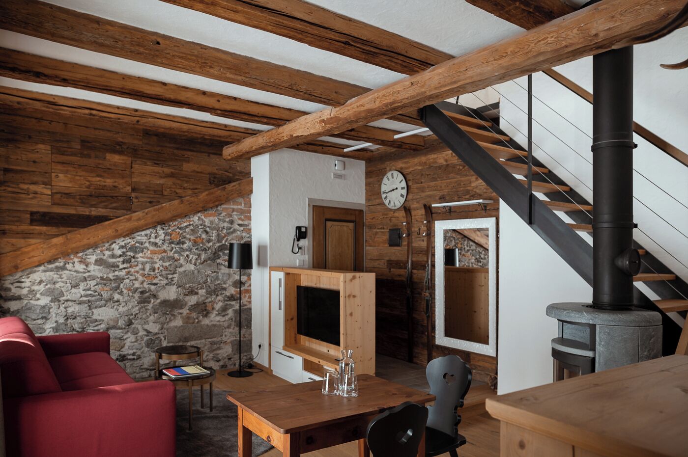Un interior rústic amb bigues de fusta al descobert, una paret de pedra, una taula de fusta amb caires i un sofà vermell, i una escala amb una canonada negra.