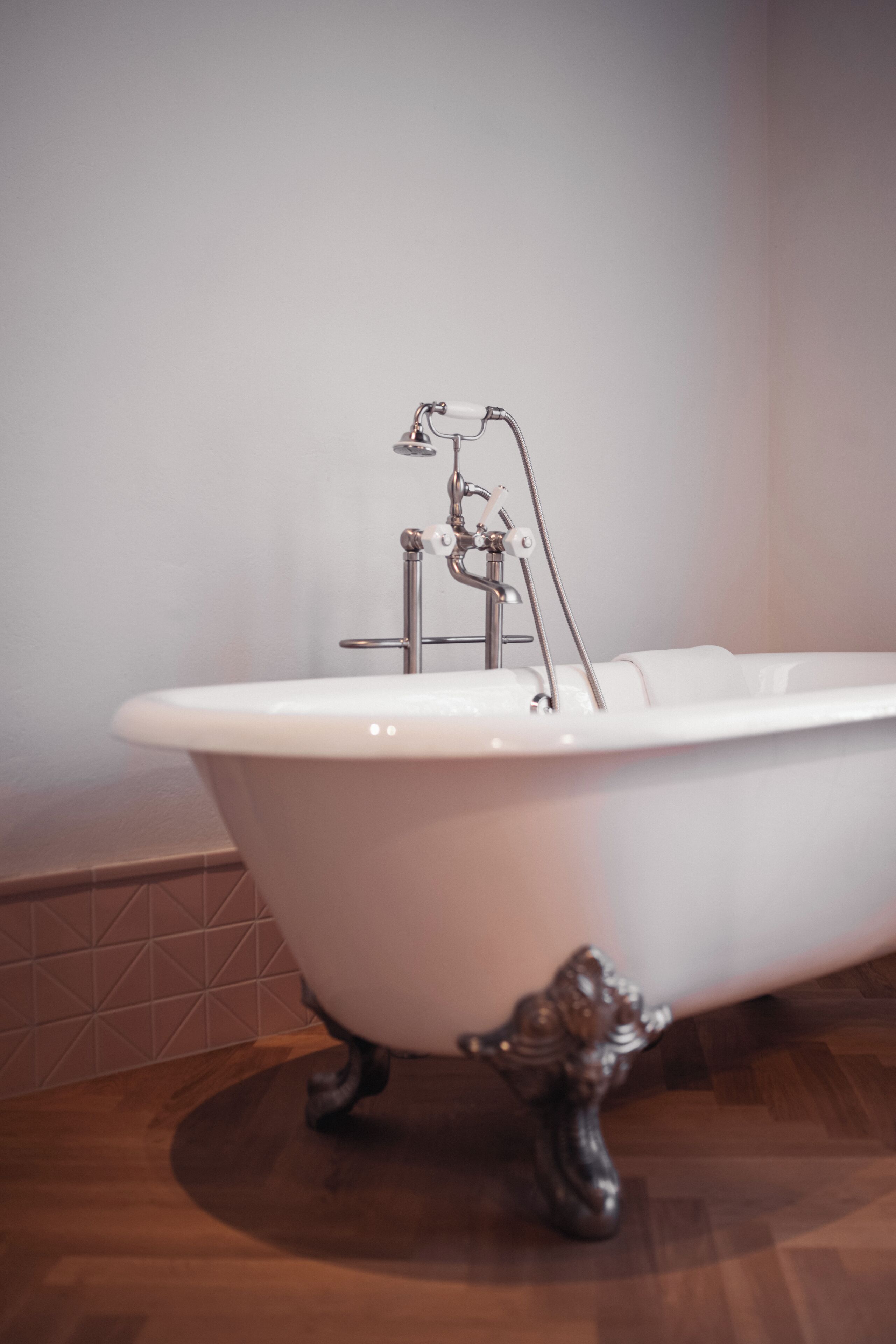 Una bañera blanca de patas de garra con un grifo y ducha antiguos, frente a una pared lisa y piso de madera.