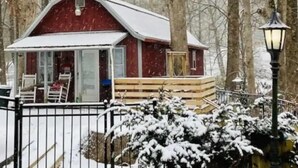 A snow-covered cabin with a porch, a wooden deck, and a black metal fence.