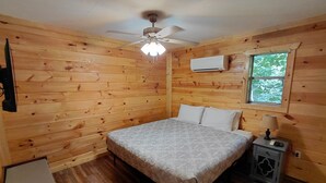 A wooden cabin bedroom with a bed, a ceiling fan, an air conditioning unit, a nightstand with a lamp, and a window with a view of trees.