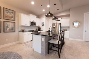 A modern kitchen with white cabinets, a central island, pendant lighting, and a dining area with chairs.