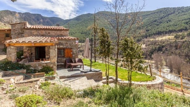A stone building with a tiled roof, a patio with outdoor furniture, and a view of a valley with mountains in the background. - Casa de Muntanya con Encanto (Arsèguel)