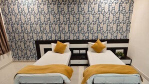 A double room with two beds, each with a white mattress and a mustard-colored blanket. The room has a patterned wallpaper and a small bedside table with a lamp.
