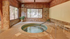 Una piscina interior con paredes de piedra, techo de madera, ventanas amplias y una escalera de metal.