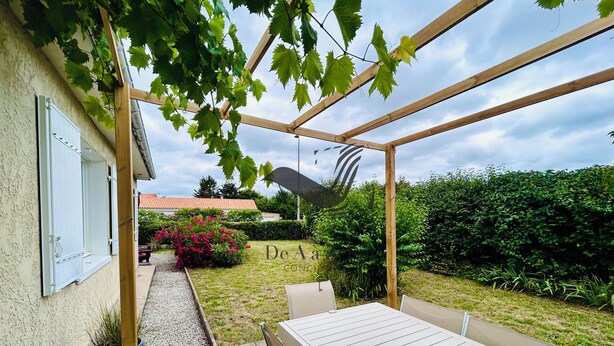 A covered patio area with a table and chairs, a pergola with climbing plants, a garden with a hedge, and a building with a window and door. - Chavid 17 - Spacieuse Maison Avec Jardin (Échillais)