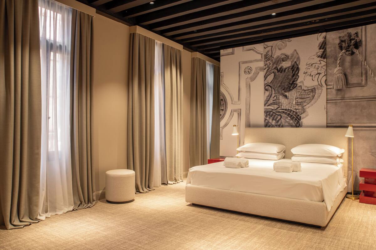 A modern hotel room with a large bed, beige curtains, a small round ottoman, and a decorative wall mural.