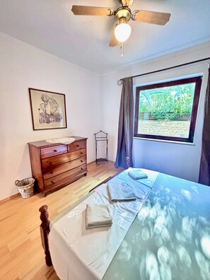 Une chambre à coucher comprenant un lit, une commode en bois, un ventilateur de plafond, une fenêtre avec des rideaux et un tableau encadré accroché au mur.