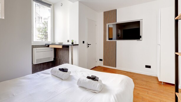 Una habitación de hotel moderna con una cama grande, un televisor montado en la pared, una kitchenette pequeña y una ventana con vistas al exterior.