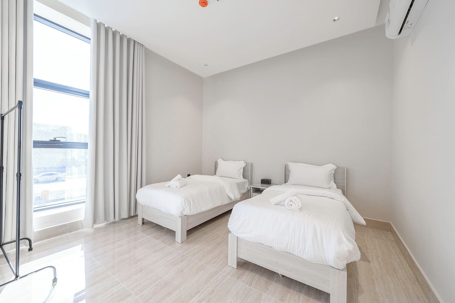A modern hotel room with two single beds, a large window with curtains, a flat-screen TV, and a ceiling-mounted air conditioner.