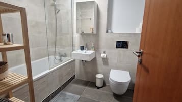 A modern bathroom with a glass shower enclosure, a white sink, a toilet, and a wooden shelf with towels.
